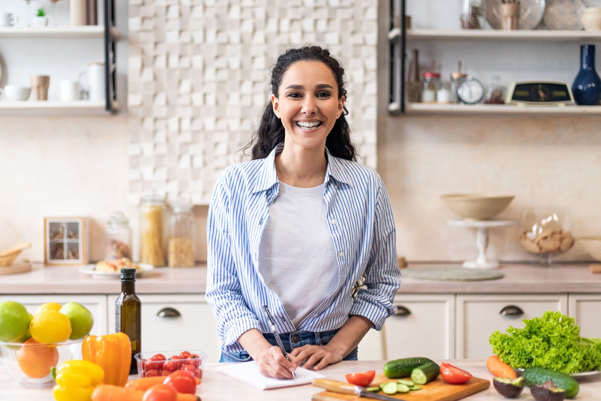 Happy housewife writing new food recipes in her notebook, standing in home kitchen interior and smiling at camera